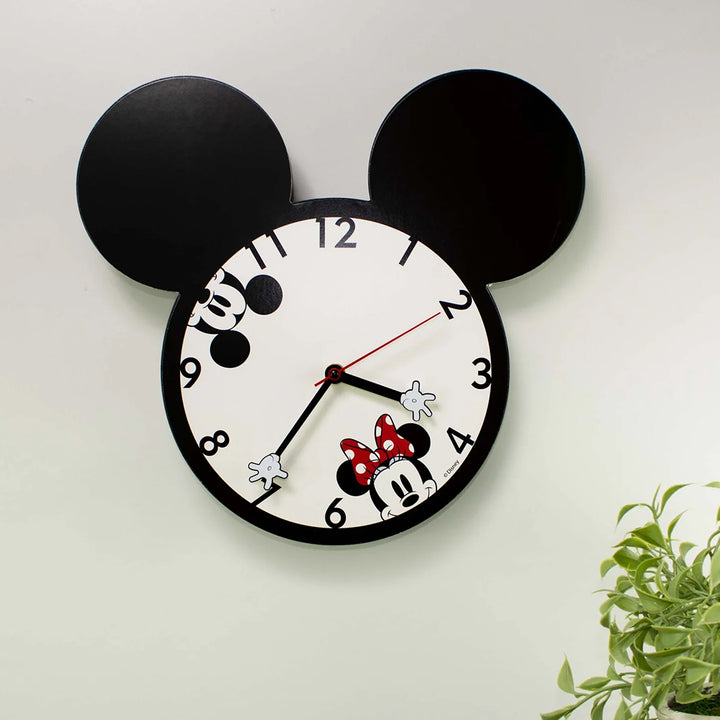 Mickey and Minnie Mouse Wall Clock With Ears mounted on white wall showing matte black Mickey ear silhouette shape and decoupage character clock face with green plant in corner officially licensed Disney wall clock by Bioworld